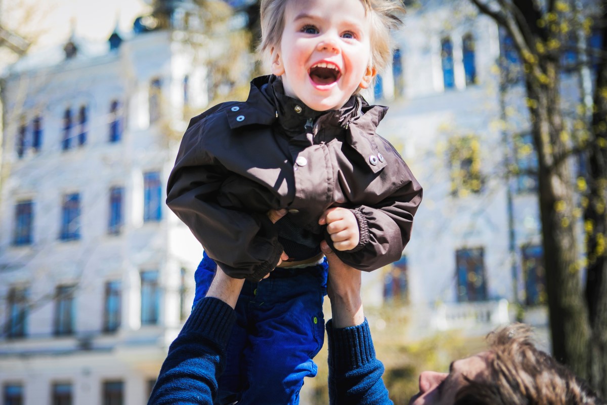 Leikki-ikäinen lapsi nauraa innoissaan, kun isä nostaa hänet korkealle ilmaan.