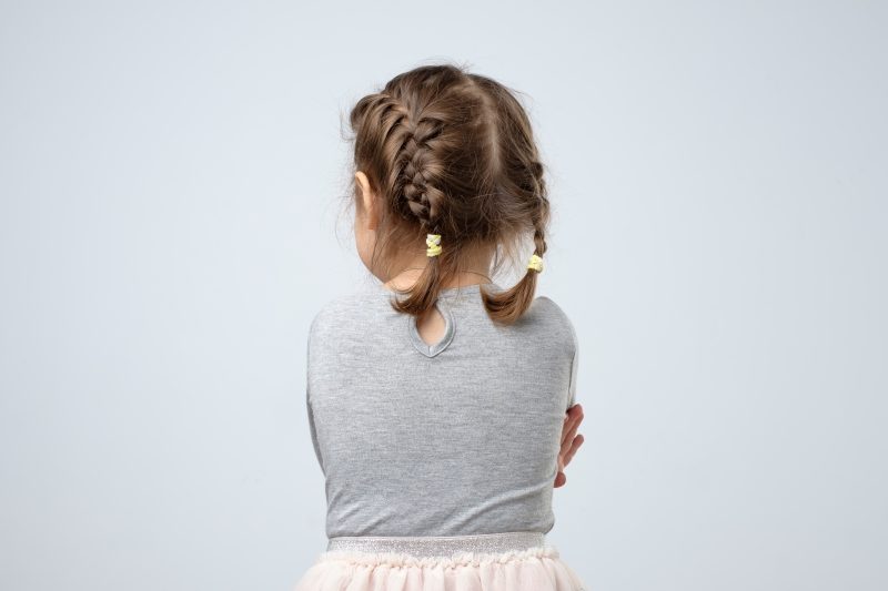 Pieni lettipäinen tyttö kuvattuna takaapäin. Omat kädet kiedottuna vartalon ympärille.