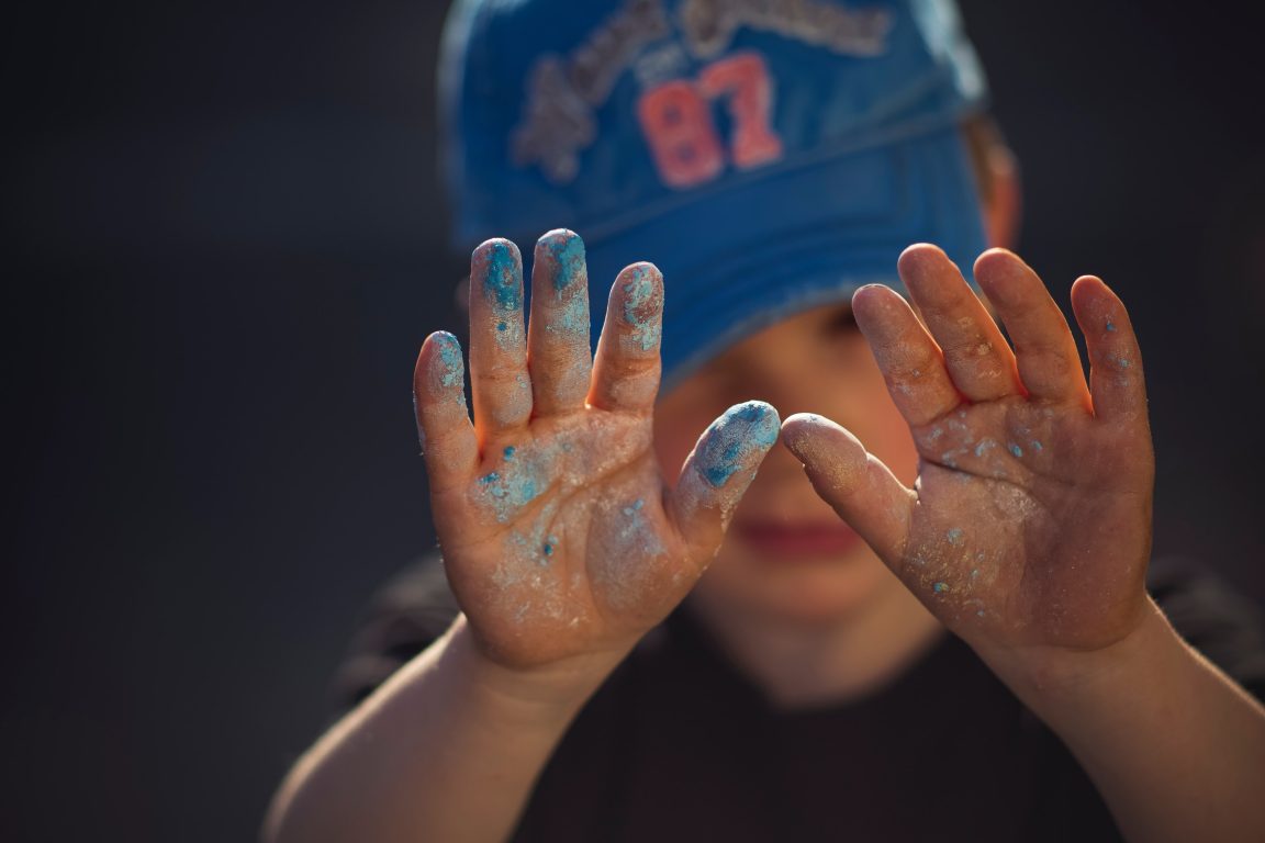 Pieni lapsi sininen lippalakki päässään näyttää sinisessä maalissa olevia käsiään kohti katsojaa.