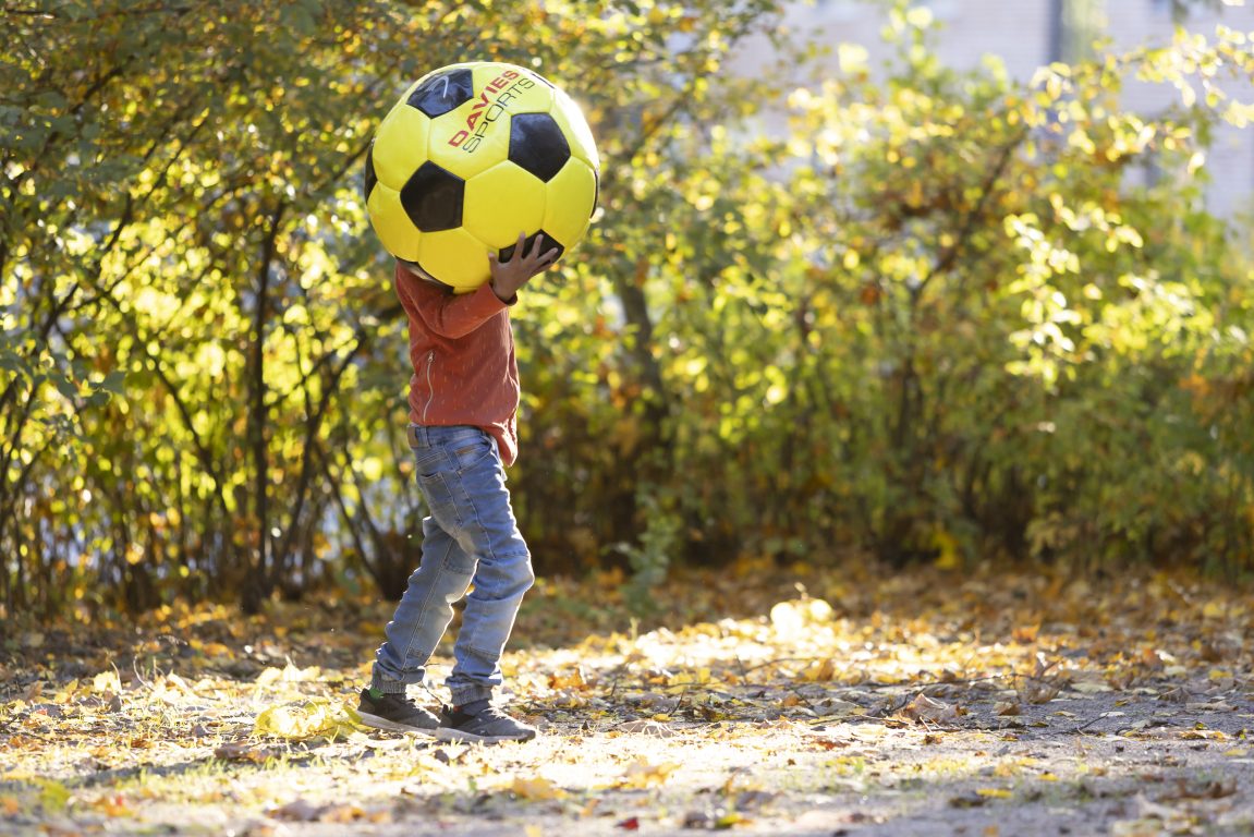 Nuori, oranssipaitainen poika leikkii ulkona isolla, keltaisella pallolla, joka peittää kuvassa hänen päänsä kokonaan.