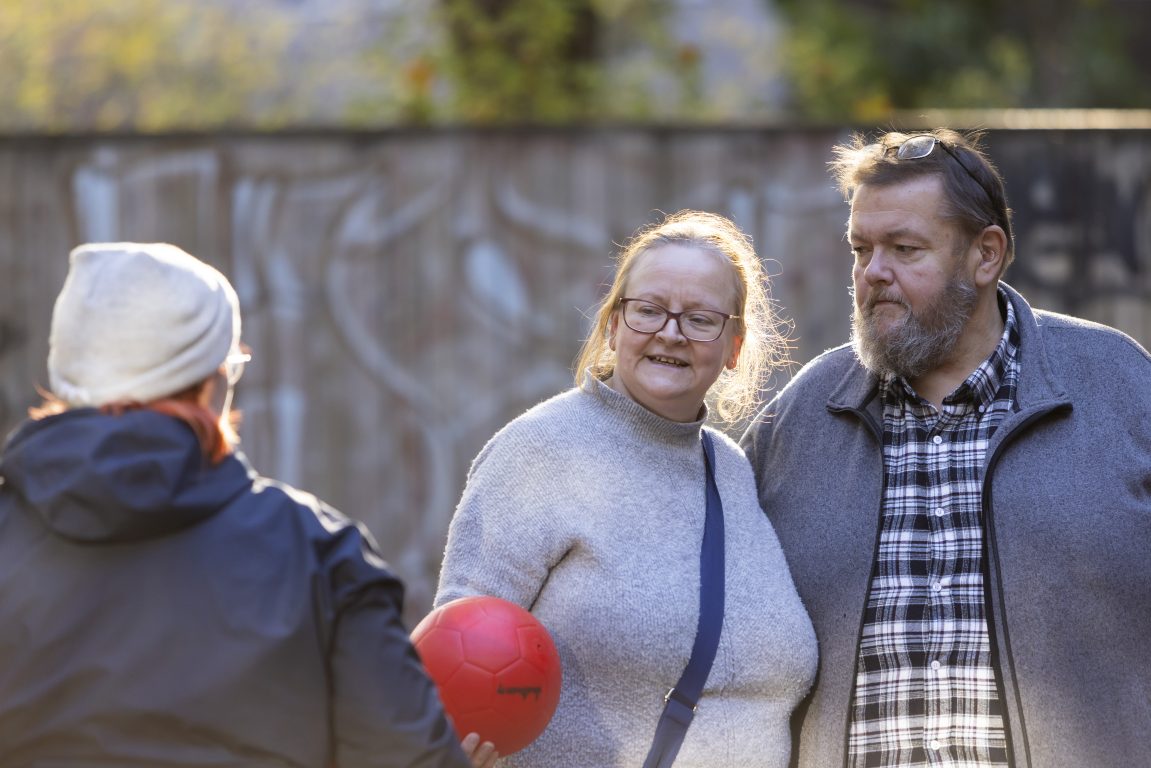 Teini-ikäinen tyttö ojentaa isälleen ja äidilleen punaisen pallon ulkona.
