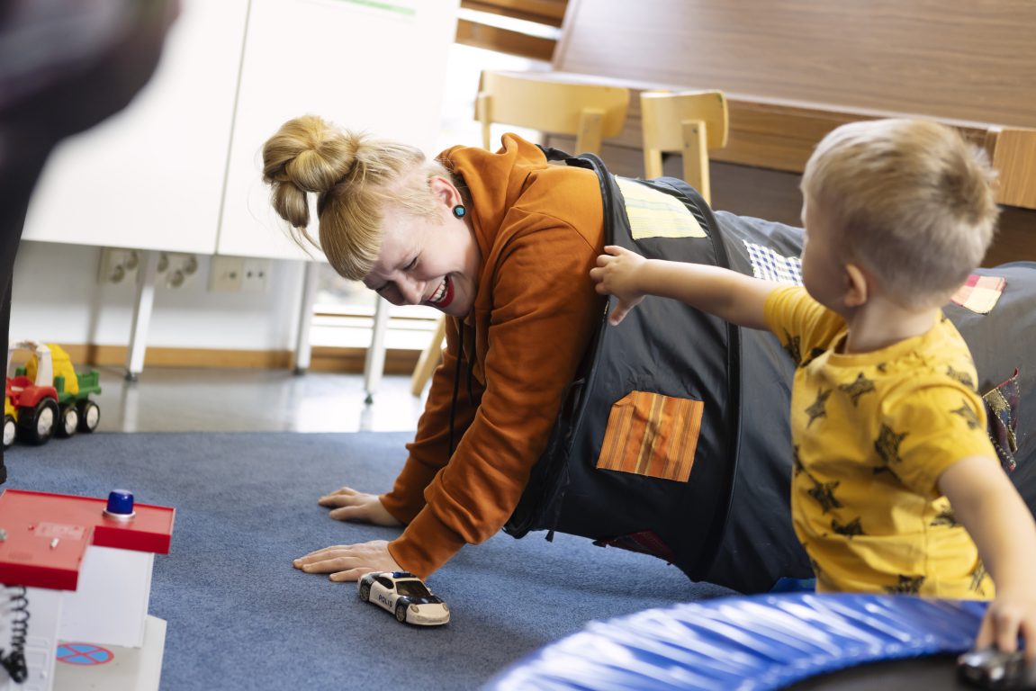 Äiti ja pikkupoika leikkivät perhekeskuksessa. Äiti on ryöminyt lasten leikkitunneliin ja jäänyt sinne jumiin. Äitiä naurattaa.