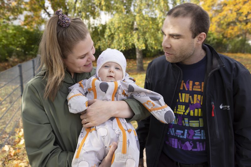 Isä, äiti ja vauva ovat ulkona syksyisessä maisemassa. Vauva on äidin sylissä ja sekä isä että äiti katsovat häntä.