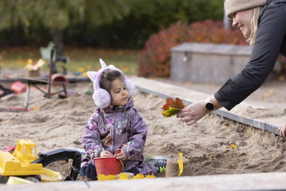 Pikkutyttö leikkii hiekkalaatikolla ja nainen ojentaa hänelle lehden.