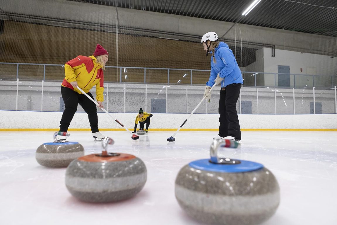 Keltatakkinen nainen ja sinitakkinen tyttö seisovat jäähallissa curlingharjojen kanssa ja taaempana keltatakkinen mies on työntämässä curlingkiveä heidän suuntaansa.