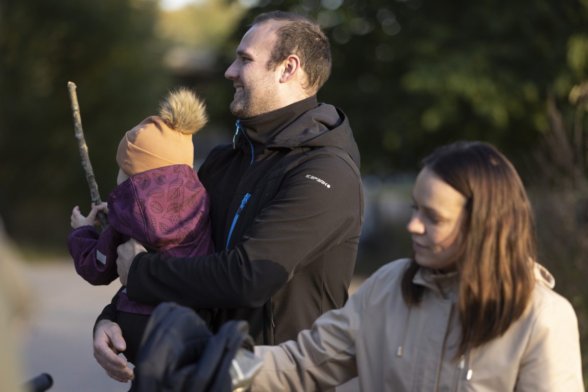 Harmaatakkinen nainen, mustatakkinen nainen ja lilatakkinen pikkutyttö ulkoilevat. Tyttö on miehen sylissä ja pitää kädessään keppiä. He katsovat poispäin kamerasta ja isä hymyilee,