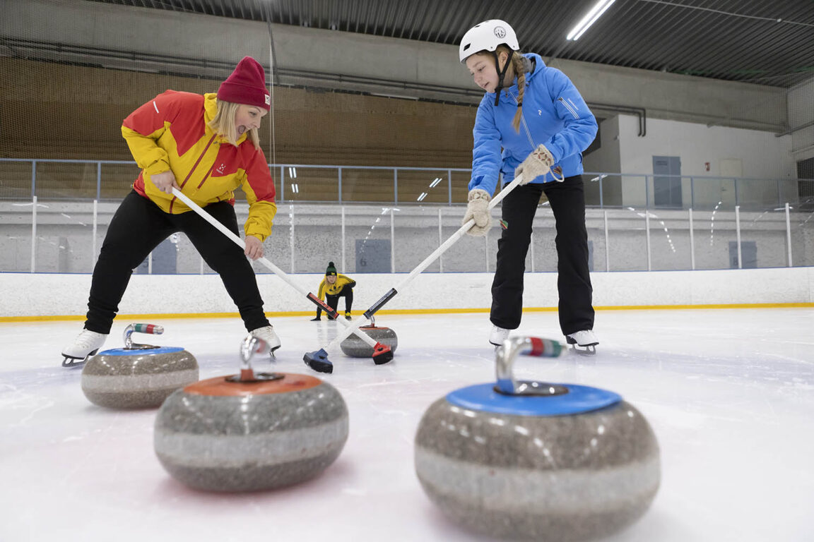 Keltatakkinen nainen ja kaksi teini-ikäistä nuorta pelaavat curlingia jäähallissa. Kaikilla on iloiset ilmeet.