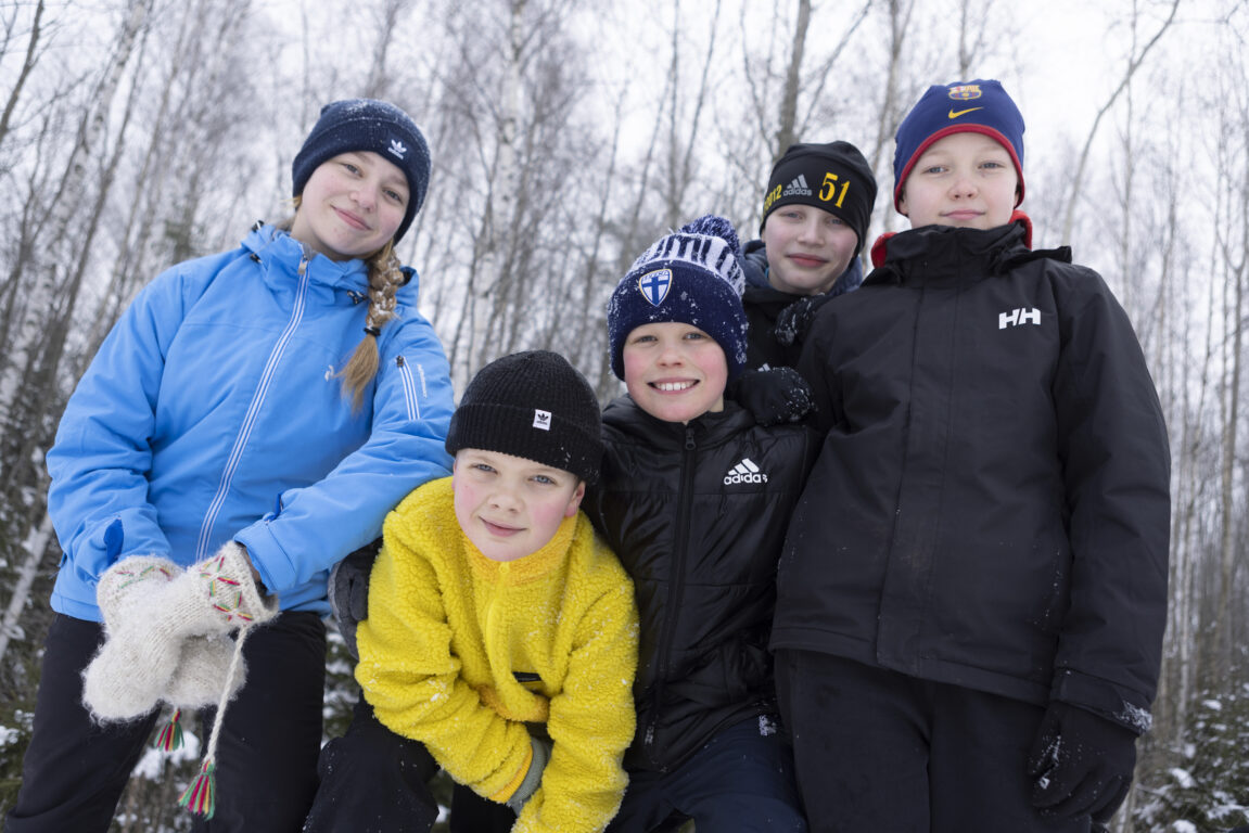 Viisi eri-ikäistä lasta seisovat ulkona lumisessa maisemassa ja katsoo kameraan.