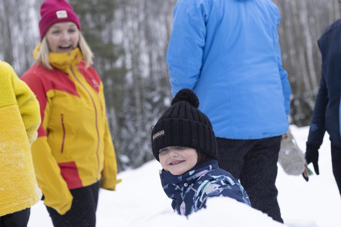 Mustapipoinen lapsi istuu hangessa ja hymyilee kameralle myssy silmillä. Taustalla näkyy hymyileviä aikuisia.