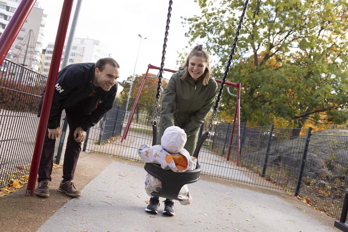 Pieni, vaaleanpunaiseen haalariin puettu lapsi keinuu vauvakeinussa. Hänen äitinsä antaa hänelle hymyillen vaihtia ja isä on kumartunut hymy kasvoillaan lapsen puoleen.